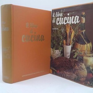 May include: A hardbound cookbook titled Il Libro di Cucina with a solid orange cover and gold lettering, displayed alongside its matching dust jacket featuring a photographic collage of kitchen items, herbs, and a wine bottle with a straw casing.