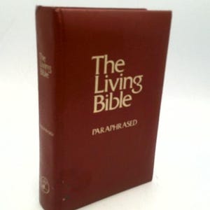 May include: A maroon-colored book titled "The Living Bible" with gold lettering. The book is paraphrased and has a simple, classic design. The book is standing upright.
