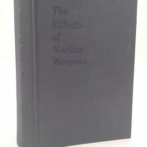 May include: A dark gray hardcover book titled The Effects of Nuclear Weapons. The front cover features the title in dark lettering, and the spine displays the text The Effects of Nuclear Weapons 1977 in a vertical orientation against the solid cover.