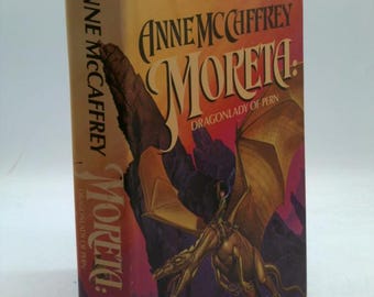 May include: A hardcover book titled "Moreta: Dragonlady of Pern" by Anne McCaffrey. The cover features a dragon with a rider, set against a backdrop of purple and gold hues. The book's spine displays the author's name and title.