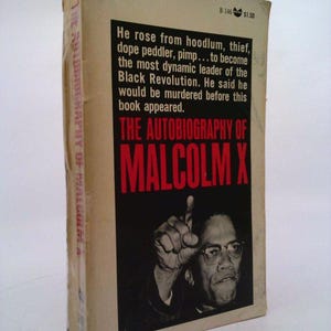 May include: A vintage paperback book cover with a black and white photo of Malcolm X. The cover is beige with red text that reads "The Autobiography of Malcolm X".