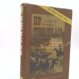 May include: A brown book titled "Up the Trail in '79" by Baylis John Fletcher. The book is part of The Western Frontier Library series and is published by the University of Oklahoma Press. The cover features a black and white illustration of a wagon train crossing a river.
