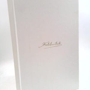 May include: A white book with the word "Fidebartek" in gold script on the cover. The book is standing upright, with the spine visible on the left side. The cover is smooth and clean.