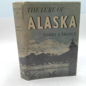 May include: Vintage book titled "The Lure of Alaska" by Harry A. Franck. The cover features a landscape illustration of mountains, a town, and a silhouette of a building. The title is in large, gold-colored letters.