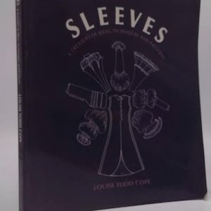 May include: A dark book titled "SLEEVES" with white text and illustrations. The cover features a drawing of various sleeve styles and the author's name, Louise Todd Cope. The book is a guide to ideal techniques and patterns.