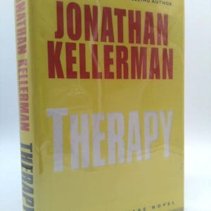 May include: A hardcover book titled "Therapy" by Jonathan Kellerman. The book has a yellow cover with the author's name in large red letters and the title in white. The text "An Alex Delaware Novel" is at the bottom.