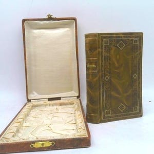 May include: An open, rectangular brown box with a cream-colored interior and a gold clasp, next to a vintage book with gold-toned cover and the word "Mineral" on the spine. The book has decorative gold accents.