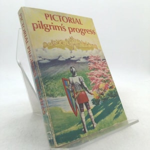 May include: A vintage book titled "Pictorial Pilgrim's Progress" is displayed on a clear acrylic stand. The cover features an illustration of a knight in armor, holding a shield, standing in a landscape with a castle and pink flowering trees.