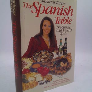May include: A hardcover cookbook titled "The Spanish Table" by Marimar Torres. The cover features a woman, various Spanish dishes, and wine bottles. The book's spine is brown with the title and author's name.
