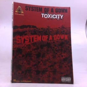 May include: A music book titled "Toxicity" by System of a Down. The cover is predominantly red with the band's name and album title in white and black. The Hal Leonard logo is visible.
