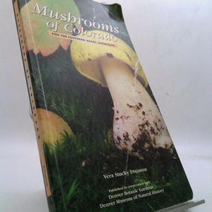 May include: A book titled "Mushrooms of Colorado and the Southern Rocky Mountains" by Vera Stucky Evenson. The cover features a close-up of a large, yellow-capped mushroom with a white stem growing in a bed of green moss.