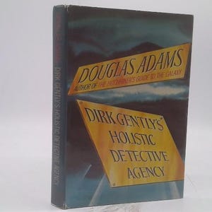 May include: A hardcover book titled "Dirk Gently's Holistic Detective Agency" by Douglas Adams. The cover features a dark blue and teal background with two yellow diamond-shaped text boxes. The spine reads "Dirk Gently's Holistic Detective Agency."