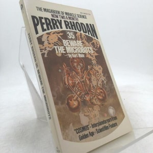 May include: A vintage paperback book titled "Perry Rhodan 35 Beware the Microbots" by Kurt Mahr. The cover features a brown and beige color scheme with an illustration of a robot on a vehicle. The book is propped up on a clear stand.