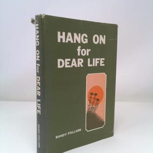 May include: A vintage book titled "HANG ON for DEAR LIFE" by Randy Pollard. The cover is dark green with white lettering and a rectangular illustration of a sunset with trees. The book's spine also features the title.