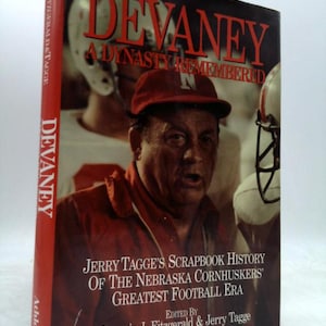 May include: A hardcover book titled "Devaney: A Dynasty Remembered" featuring a portrait of a man in a red hat. The book's cover includes text about the Nebraska Cornhuskers' football era. The spine displays the title and the publisher's name.