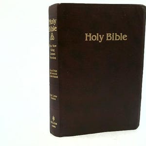 May include: A brown leather-bound Holy Bible with gold lettering. The title "Holy Bible" is prominently displayed on the cover. The spine includes text indicating the version and publisher. The book is standing upright.