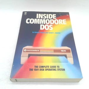 May include: A vintage book titled "INSIDE COMMODORE DOS" with a rainbow graphic and a Commodore 1541 disk drive illustration. The book's cover is blue with white text and the subtitle "THE COMPLETE GUIDE TO THE 1541 DISK OPERATING SYSTEM."
