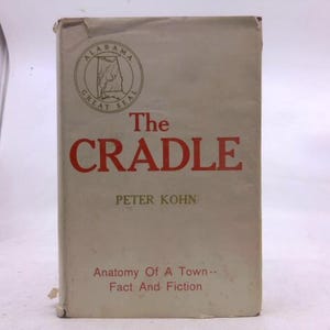 May include: A vintage book titled "The Cradle" by Peter Kohn. The cover is off-white with red lettering and features the Alabama Great Seal. The book's subtitle reads "Anatomy Of A Town--Fact And Fiction."