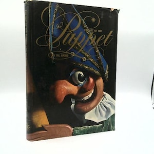 May include: A hardcover book titled "The Art of the Puppet" by Bil Baird. The cover features a colorful illustration of a puppet's face with a large nose, wide eyes, and a blue hat with gold trim. The book is set against a dark background.