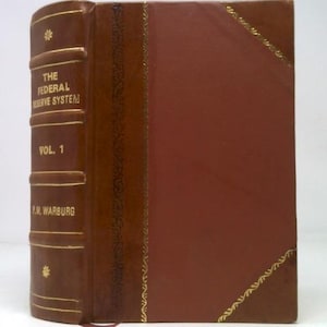 May include: A vintage book titled "The Federal Reserve System, Vol. 1" by P.M. Warburg. The book has a brown leather spine with gold lettering and decorative accents. The cover is a darker shade of brown with gold trim.