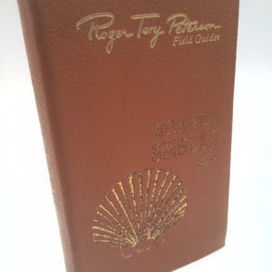 May include: A brown field guide titled "Atlantic Shells" by Roger Tory Peterson. The cover features gold lettering and a gold seashell illustration. The book is a guide for identifying shells.