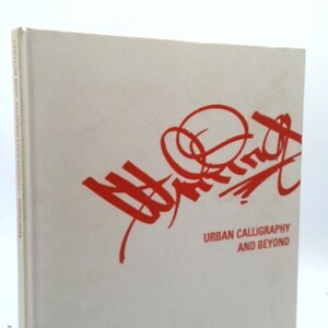 May include: A hardcover book titled "Urban Calligraphy and Beyond." The book has a light gray cover with red stylized lettering and the title in red. The book is standing upright.