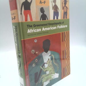 May include: A hardcover book titled The Greenwood Encyclopedia of African American Folklore, Volume 1 A-F, edited by Anand Prahlad. The cover features a green and red illustration of a person holding a green striped cat and a pipe.