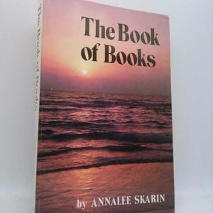 May include: A hardcover book titled "The Book of Books" with a sunset over the ocean on the cover. The cover is predominantly shades of orange, pink, and purple. The author's name, Annalee Skarin, is printed at the bottom.