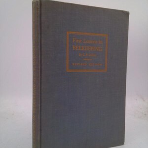 May include: A hardcover book titled "First Lessons in Beekeeping" by C.P. Dadant. The book has a gray cover with orange text and a rectangular border. The spine of the book also has text. The book is a revised edition.