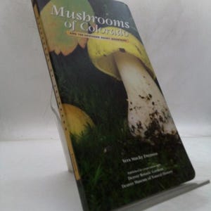 May include: A hardcover book titled "Mushrooms of Colorado" with a photograph of a mushroom on the cover. The book is propped up on a clear stand. The cover features green, yellow, and white colors.