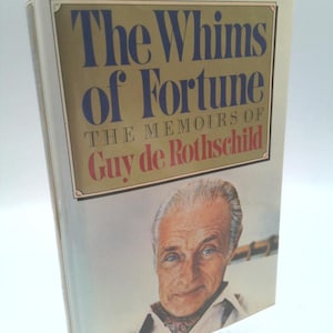 May include: A hardcover book titled "The Whims of Fortune: The Memoirs of Guy de Rothschild." The cover features a portrait of a man with blue eyes and a white shirt. The title is in blue and the author's name is in red.