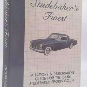 May include: A hardcover book titled "Studebaker's Finest" with a dark blue spine and a light beige cover. The cover features a drawing of a dark blue Studebaker car and the text "A History & Restoration Guide for the '53-'54 Studebaker Sports Coupe."