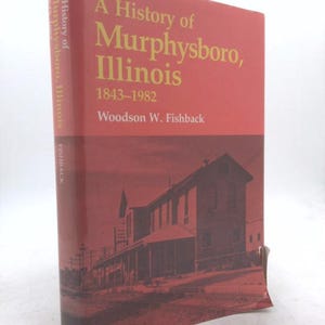 May include: A hardcover book titled "A History of Murphysboro, Illinois 1843-1982" by Woodson W. Fishback. The book has a red cover with gold lettering and a sepia-toned image of a building. The spine reads "A History of Murphysboro, Illinois".