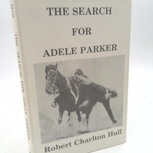 May include: A hardcover book titled "THE SEARCH FOR ADELE PARKER" by Robert Charlton Hull. The cover is off-white with black text and a black and white image of a horse and rider. The book's spine also has the title.