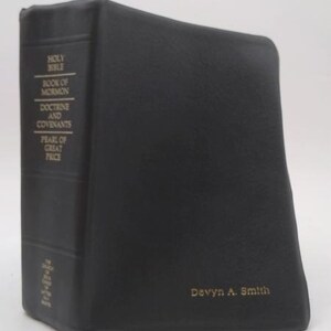 May include: A black leather-bound book featuring gold embossed text on the spine that reads Holy Bible, Book of Mormon, Doctrine and Covenants, and Pearl of Great Price. The name Devyn A. Smith is embossed in gold on the front cover of the book.