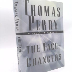 May include: A hardcover book titled "The Face-Changers" by Thomas Perry. The book has a light gray cover with the title and author's name in dark gray. The book is a novel of suspense.