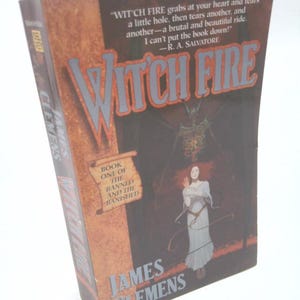 May include: A hardcover book titled "Witch Fire" by James Clemens. The cover features a dark, illustrated scene with a woman in a white dress and the book's title in large, stylized letters. The book is part of "The Banned and the Banished" series.