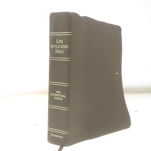 May include: A brown leather-bound Bible with gold lettering. The spine reads "Life Application Bible" and "New International Version." The publisher's name, Zondervan, is printed at the bottom of the spine. The book is closed and angled slightly.