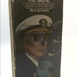 May include: A book cover with a dark background featuring a man in a navy blue uniform holding a globe. The title of the book is "The Brink" and the author is "Rick Setlowe".
