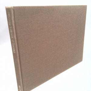 May include: A hardcover book with a brown cover. The book's spine features the title "William Kurelek O'Toronto" and the publisher "new press" in white text. The book is angled, showcasing its textured cover.