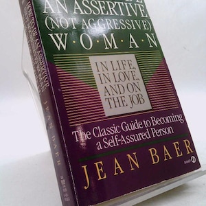May include: A purple and green book titled "How to Be an Assertive (Not Aggressive) Woman" by Jean Baer. The book cover features the text "In Life, In Love, and on the Job" and "The Classic Guide to Becoming a Self-Assured Person".