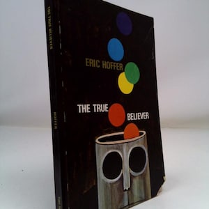 May include: A hardcover book titled "THE TRUE BELIEVER" by Eric Hoffer. The cover is black with colorful circles and a stylized illustration of a face. The book's spine reads "THE TRUE BELIEVER" and "HOFFER".