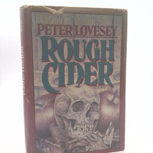 May include: A hardcover book titled "Rough Cider" by Peter Lovesey. The cover features a skull, apples, and the title in large, teal letters. The book has a brown spine and a worn appearance.