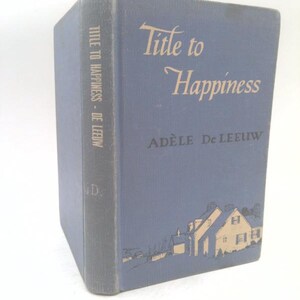 May include: A blue hardcover book titled Title to Happiness by Adèle De Leeuw. The front cover features a cream-colored illustration of a house at the bottom. The spine displays the title and author name in white text against the blue fabric cover.