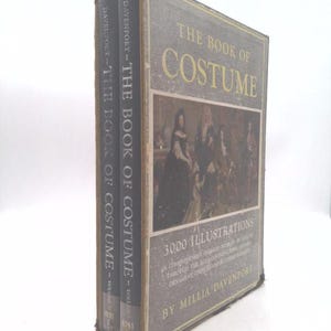 May include: A vintage book titled "The Book of Costume" with two volumes. The cover is gray with gold lettering and features an illustration of people in historical attire. The book includes 3000 illustrations and is by Millia Davenport.