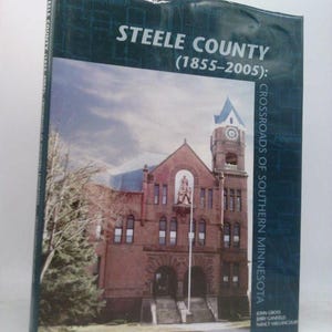 May include: A hardcover book titled "Steele County (1855-2005): Crossroads of Southern Minnesota." The cover features a photograph of a brick building with a clock tower and a blue sky. The book is wrapped in clear plastic.