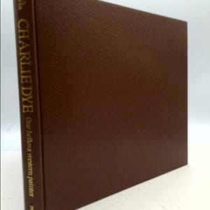 May include: A brown leather-bound book with gold lettering on the spine. The title is "Charlie Dye: One helluva western painter" by Pat Weaver.