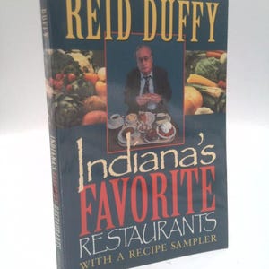 May include: A hardcover book titled Reid Duffy Indiana's Favorite Restaurants With A Recipe Sampler. The dark blue cover features a central photograph of a person sitting at a table with various dishes, flanked by images of fresh produce.