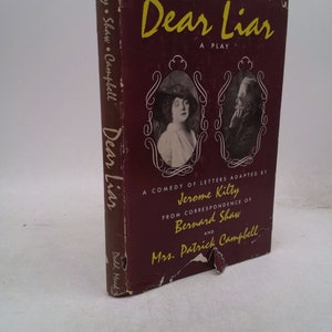 May include: A burgundy book cover with the title "Dear Liar" in yellow lettering. The cover features a black and white photo of a woman and a man. The text below the title reads "A comedy of letters adapted by Jerome Kilty from correspondence of Bernard Shaw and Mrs. Patrick Campbell."