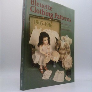 May include: A hardcover book titled "Bleuette Clothing Patterns 1905-1916" with a green cover. The cover features a doll in a dress, along with other doll clothing and accessories. The book's spine reads "Bleuette Clothing Patterns 1905-1916".